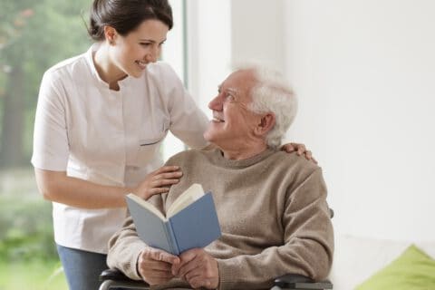 Symbolbild einer Pflegekraft, die sich um einen Heimbewohner kümmert, der ein Buch liest