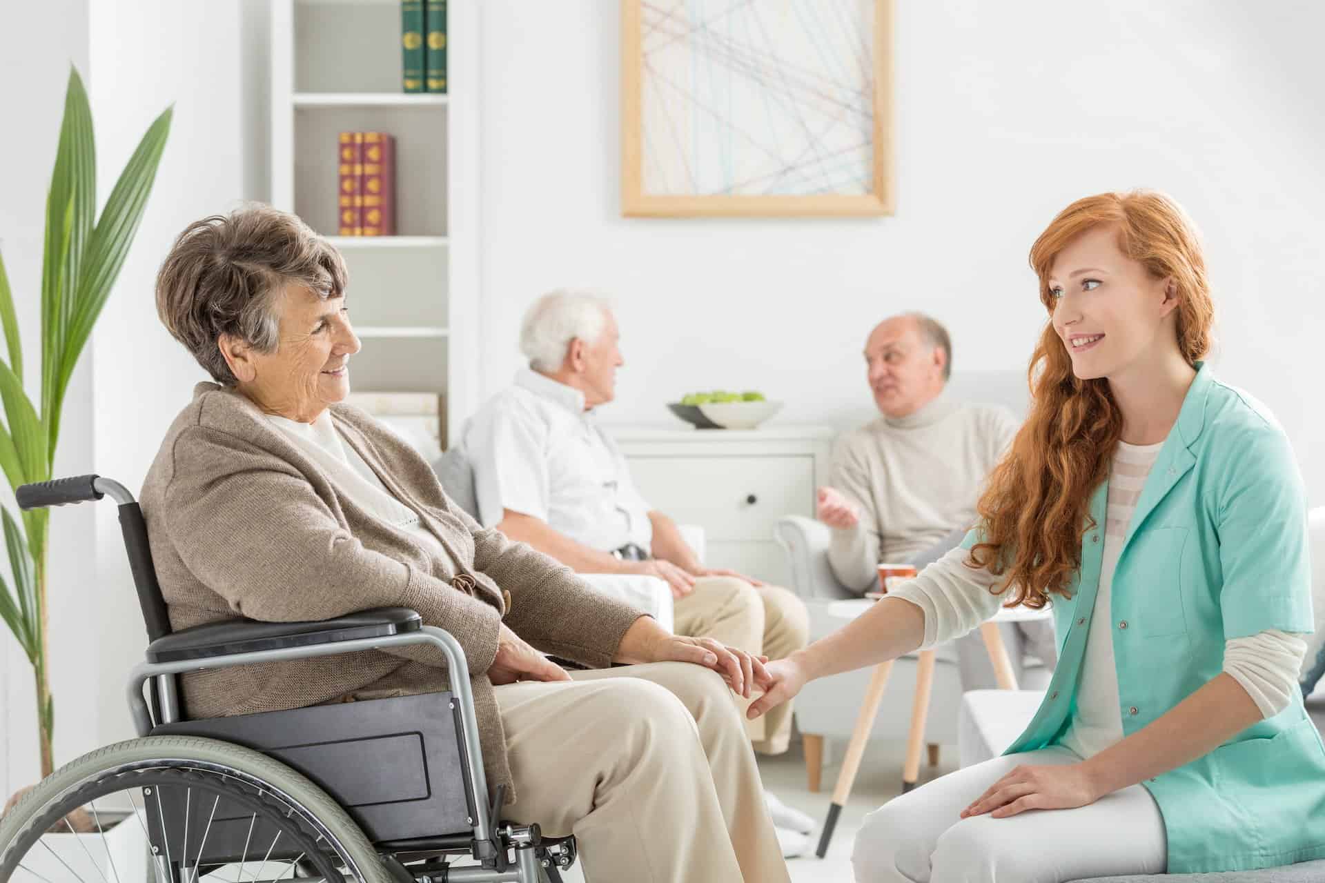 Symbolfoto einer Pflegefachkraft, die die Hand einer Heimbewohnerin im Rollstuhl hält und ihr zulächelt